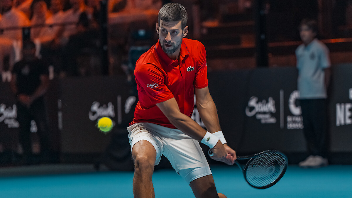 Male tennis player in red shirt and white shorts hitting a backhand shot on an indoor hard court, with audience and event signage visible in the background.