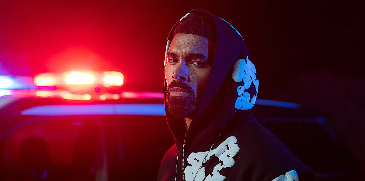 Man in a black hoodie with white patterns standing outdoors at night in front of a police car with red and blue flashing lights, creating a dramatic and intense atmosphere.