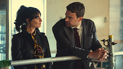 A woman in a black jacket and scarf and a man in a suit lean on a railing indoors, facing each other and having a serious conversation in a sunlit room.