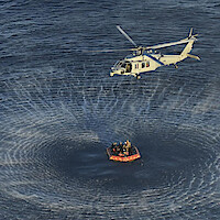 Helicopter hovering above ocean, creating circular ripples, while a group of people wait in an orange life raft below, possibly during a rescue or training operation at sea.