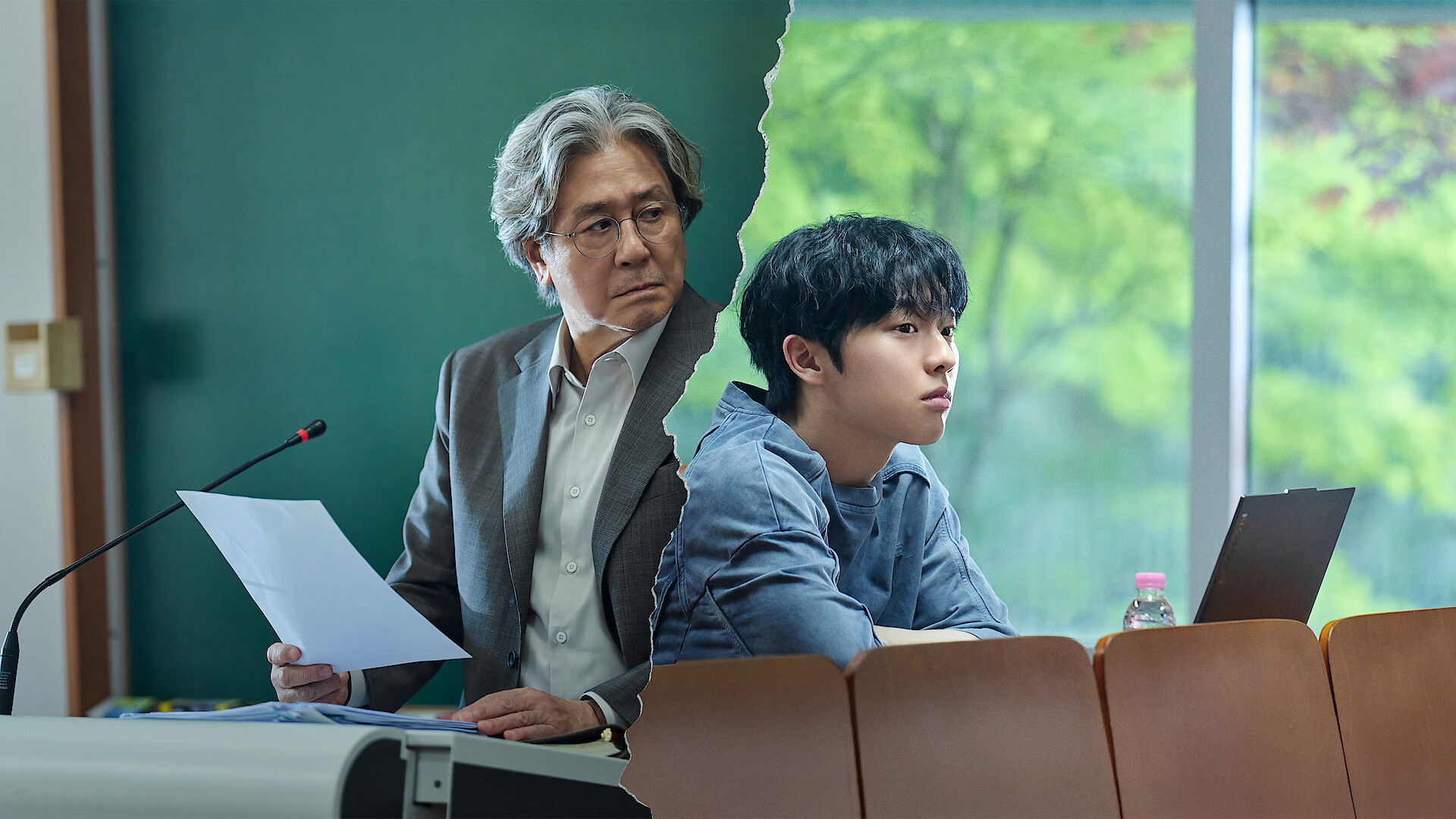 An older man in a suit holds papers at a podium in a classroom, while a young man sits at a desk with a laptop, divided by a jagged line, suggesting a tense or dramatic situation in an academic setting.