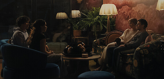Charles Melton as Austin Davis, Cailee Spaeny as Ashley Miller, Carey Mulligan as Lindsay Crane-Martin, and Oscar Isaac as Josh Martin  sit in pairs on sofas in a dimly lit, cozy living room with vintage decor, plush seating, and a coffee table.