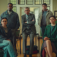 Five people, three seated and two standing, pose indoors in a well-lit room with wooden floors and stained-glass windows, dressed in formal and casual clothing, appearing serious and focused.