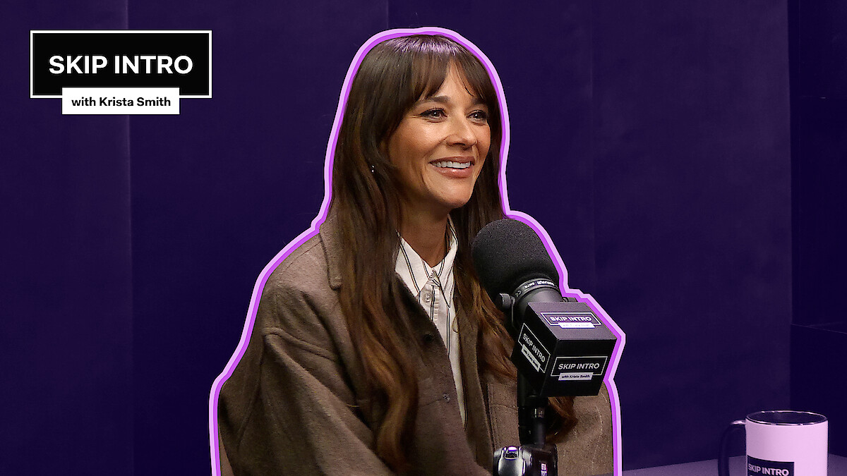 Rashida Jones, smiling, seated in front of a microphone with a dark purple background.