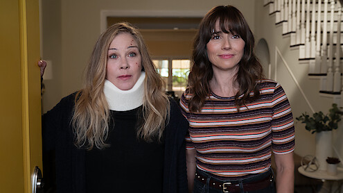 Two women standing in the entryway of a house; one wears a neck brace and dark sweater, the other wears a striped shirt. They stand near an open yellow door, with a staircase and potted plant visible in the background.