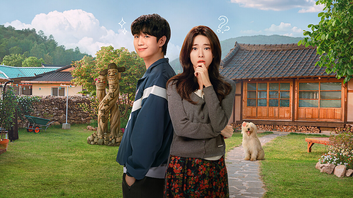 A young man and woman stand in a sunny, rural yard with traditional wooden house, lawn, garden statues, and a fluffy dog, mountains in the background, blue sky with clouds, both subjects displaying thoughtful and playful expressions.
