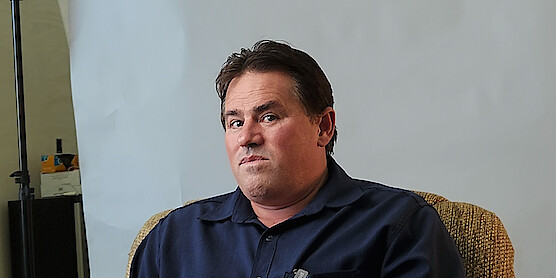 A man with brown hair in a blue collared shirt sits in front of a white background and looks directly to camera.