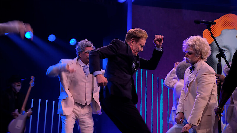 Conan O'Brien dances on stage with people dressed as Mark Twain