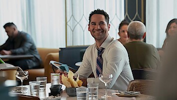 Smiling man in dress shirt and tie sitting at a restaurant table with chips, water glasses, and wine glasses, with other people dining in the background. Bright, modern interior with natural light.