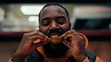 Man enjoying a piece of fried chicken indoors, with eyes closed, savoring the food in a warmly lit restaurant or fast food setting.