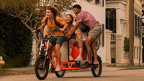 Four happy young adults riding together on a large electric bike through a sunny residential neighborhood, smiling and laughing as they share the ride on a tree-lined street with houses in the background.