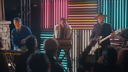 A band performs live on stage with colorful neon lights in the background, including a keyboard player, singer, and guitarist. Audience members are watching and raising their hands in an outdoor nighttime concert setting.
