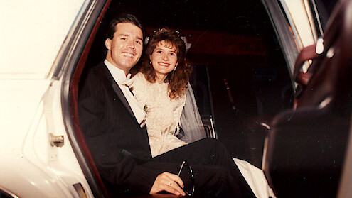 A smiling bride and groom in formal wedding attire sit together in the back seat of a car, looking joyful and relaxed, suggesting a celebratory and happy atmosphere.