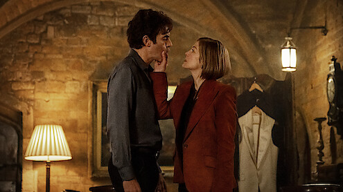 A woman in a red blazer touches a man's face in a dimly lit, stone-walled room with warm lighting, an arched ceiling, lamp, and a suit on display in the background, suggesting an intimate or dramatic interaction.