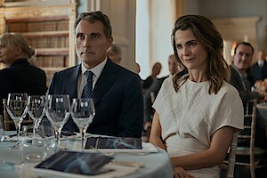 Em um ambiente formal, Keri Russell e Rufus Sewell sentam lado a lado à mesa.
