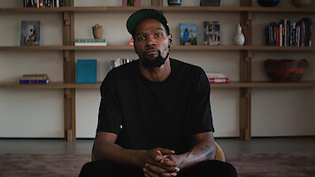 A man in a black shirt and cap sits indoors with hands clasped, in front of a bookshelf filled with books, vases, and decorative objects, giving a calm, introspective mood.
