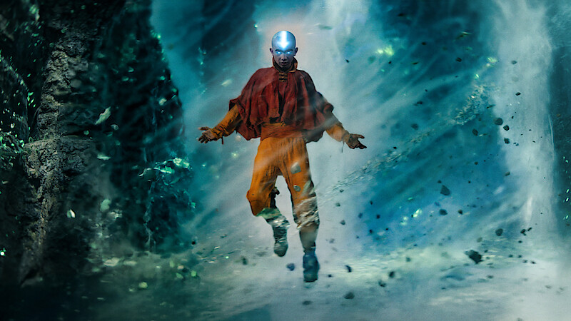 Gordon Cormier as Aang in traditional orange and red robes hovers in the air with glowing light on their head, surrounded by swirling wind and floating debris in a mystical cave or cavern environment.