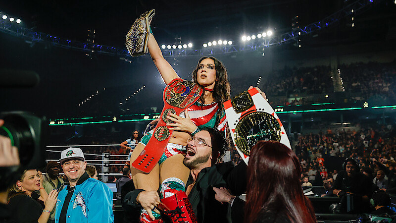 Female wrestler in a brightly lit arena celebrates, holding championship belts, surrounded by cheering fans and photographers. The mood is energetic and triumphant with a large audience in the stadium background.