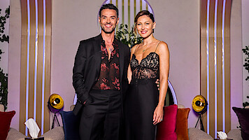 A man in a black suit and a woman in a black lace dress stand together smiling in a modern lounge with colorful cushions, round lights, plants, and decorative vertical lines in the background.