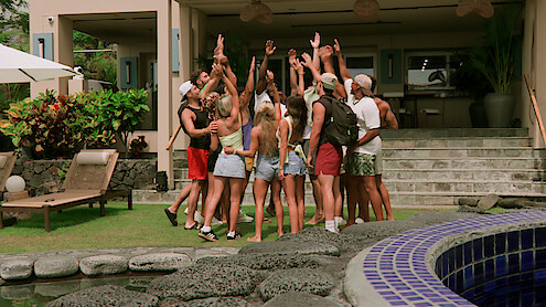 Peter Cipriano, Braeden Holliday, Scarlett Bentley, Shyanne Blankenship, Kaylee Needham, Bradley Bobbit, and Clint Smith in summer clothes stand in a circle with raised hands outside a modern villa, surrounded by greenery, lounge chairs, and a pool.