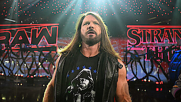 AJ Styles stands in a dramatic wrestling arena, wearing a black vest and T-shirt, with bright red RAW and STRANGER THINGS signs and stage lights in the background.