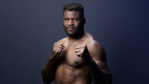 Francis Zavier Ngannou in a fighting stance with clenched fists, looking serious, posing against a plain dark gradient background in a studio setting.
