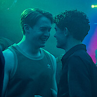 Two young men smiling closely at each other in a dimly lit nightclub with blue and green lighting, suggesting a lively social atmosphere.