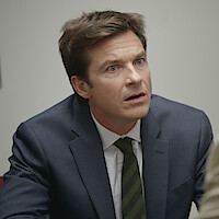 A man in a navy suit and striped tie sits indoors at a table, looking surprised or confused, in what appears to be an office or meeting room with neutral walls.