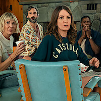 Four people sitting in a cozy wood-paneled room with a stone fireplace, all looking surprised or concerned; one woman holds a cup, another wears a “Catskills” sweater, giving the scene a tense, dramatic feeling.