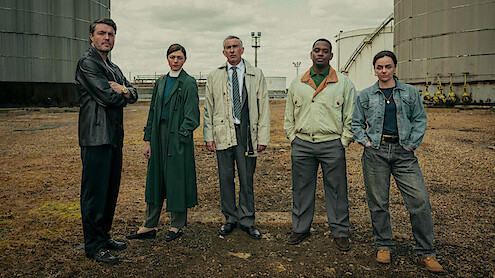 Five people stand in a gritty industrial outdoor setting between large storage tanks, dressed in various jackets and coats, facing the camera on a cloudy day, suggesting a serious or dramatic atmosphere.