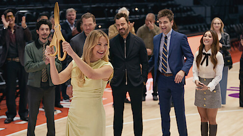 A woman in a yellow dress holds giant scissors preparing to cut a ribbon on a basketball court, surrounded by smiling, cheering people in formal and semi-formal attire during a ceremony or event.