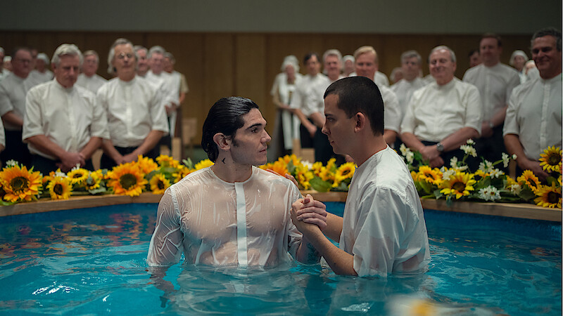 Two men in white shirts stand in a baptismal pool surrounded by sunflowers, with a group of people in white shirts and blouses watching in the background, in a brightly lit indoor setting.