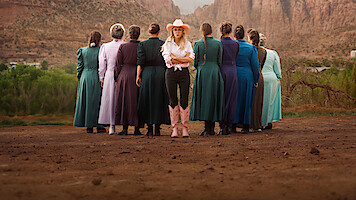 A group of women in long, modest dresses stand in a dirt area, backs to the camera, with one woman in a cowboy hat, white shirt, black pants, and pink boots facing forward against a backdrop of mountains and greenery.