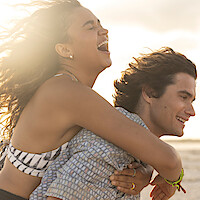 A joyful young woman laughs while riding piggyback on a smiling young man at the beach during sunset, with sunlight creating a warm, cheerful atmosphere.