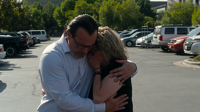 Two people embrace emotionally in a parking lot surrounded by cars and trees, suggesting comfort or consolation in a public, outdoor setting on a sunny day.