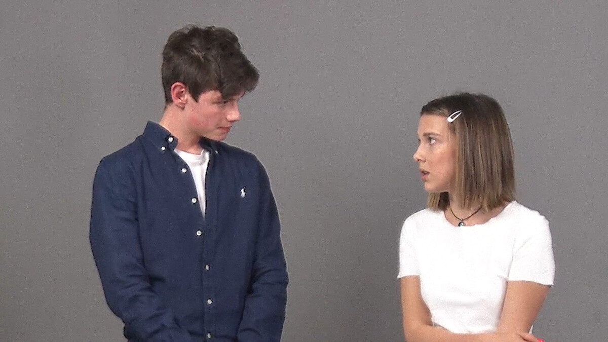 A young man in a navy shirt and a young woman in a white top stand indoors against a plain gray background, facing each other and appearing to have a conversation.