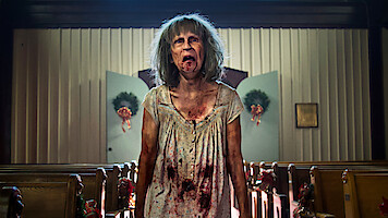 A disheveled, bloodied woman with gray hair stands in the aisle of a decorated church, with pews on either side and wreaths on the doors in the background, looking distressed or possibly zombified.
