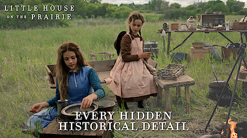 Two girls in prairie dresses sit outdoors beside rustic wooden tables and vintage cooking tools in a grassy field, reenacting a historical pioneer camp scene. A campfire is visible in the foreground, trees in the background.