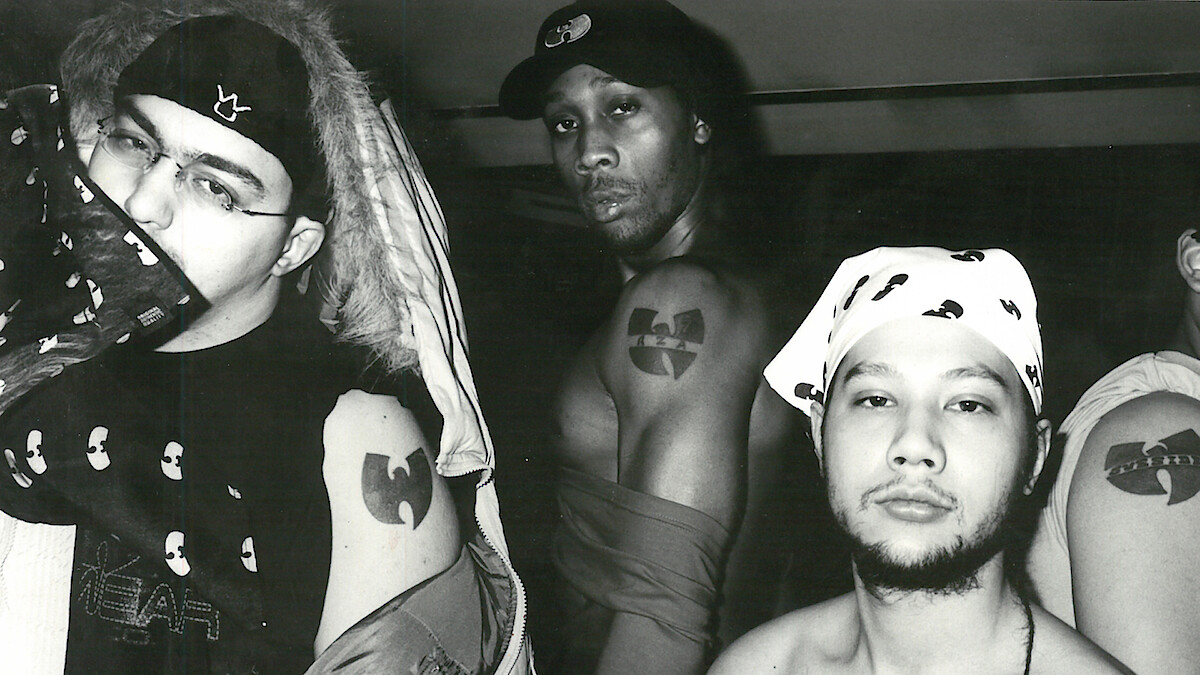 Three men pose in a black and white indoor photo, showing off Wu-Tang Clan tattoos and clothing featuring the group's logo. The environment is casual with urban streetwear and bandanas, emphasizing their affiliation and attitude.