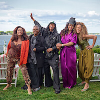 Five women posing and smiling outdoors by a lake, with Adirondack chairs on grass, all wearing colorful, expressive outfits under a partly cloudy sky.