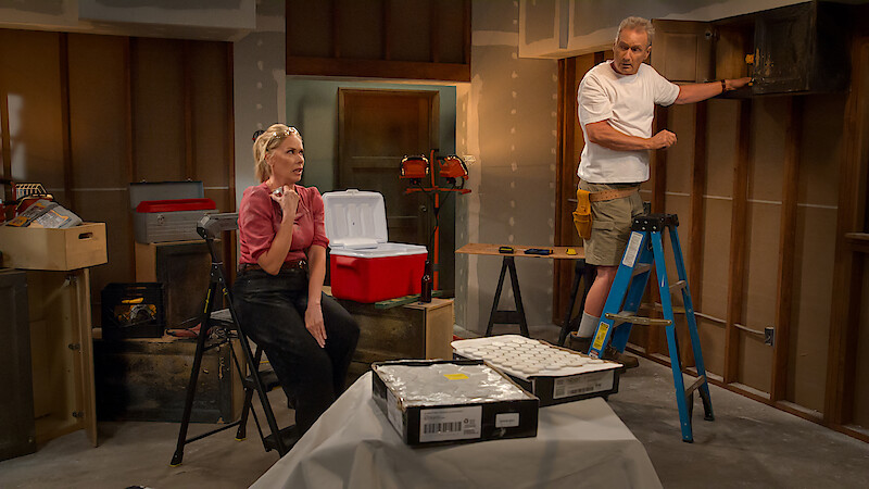 Woman sitting and man standing on ladder in unfinished garage or basement, surrounded by tools, coolers, and boxes, with exposed walls and construction materials, appearing to be in the middle of a home renovation project.