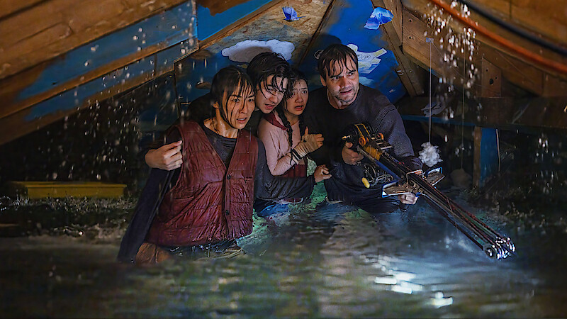 Four people waist-deep in floodwater in an attic, huddled together, appearing worried and alert, with one person holding a weapon, water pouring from ceiling, scene is tense and dramatic.