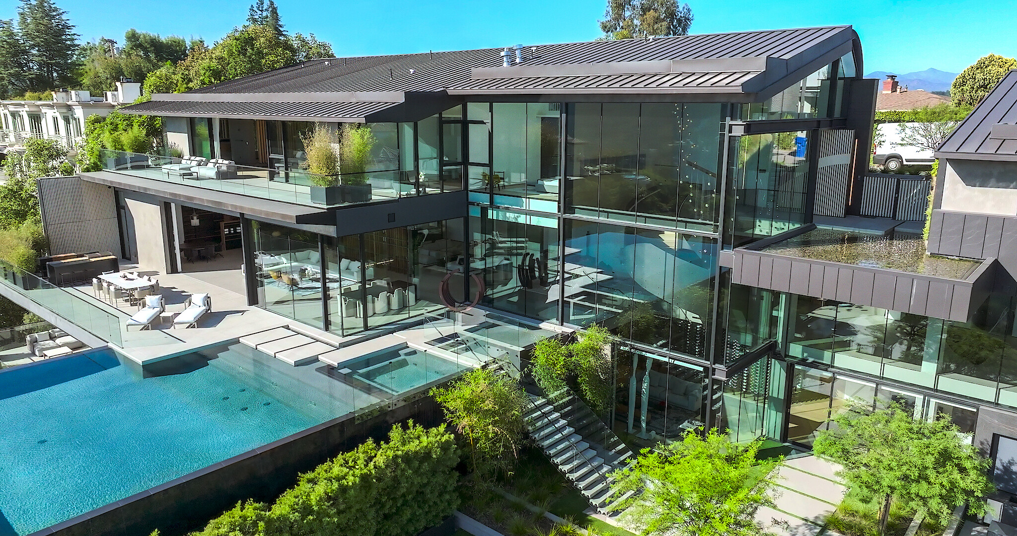 Buying Beverly Hills key art- A photo of a mansion with large glass windows, a pool, in ground jacuzzi, and stairs leading down to a garden area.