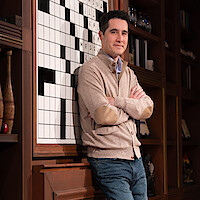 Man in casual clothes stands with arms crossed, leaning against a large crossword puzzle in a room with dark wooden bookshelves filled with books, decor, and vases. Warm, indoor lighting.