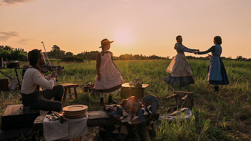 People in pioneer-era clothing gather outdoors at sunset; a man plays a violin while three women dance and stand in a grassy field with rustic tables and wooden benches, creating a nostalgic, rural atmosphere.