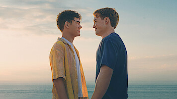 Two young men stand facing each other, smiling, at the beach during sunset with the ocean and sky in the background.