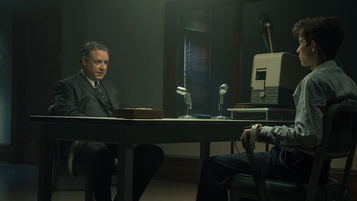 Two men sit across from each other in a dimly lit interrogation room, one appearing to question the other who is attached to a polygraph machine, with microphones and recording equipment on the table.