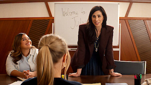 Three women in a meeting room, one standing and smiling while leaning on a table, two seated, with a whiteboard in the background that says "Welcome to". Professional office setting.