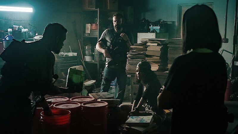Four people in a dimly lit, cluttered room filled with equipment, red buckets, and stacks of paper, appearing to be discussing or planning something in a tense, secretive environment.