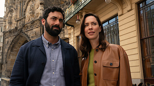 A man and woman stand outdoors in a city setting, with historic stone building details and modern architecture visible behind them. Both appear thoughtful, dressed in casual jackets, under daylight.